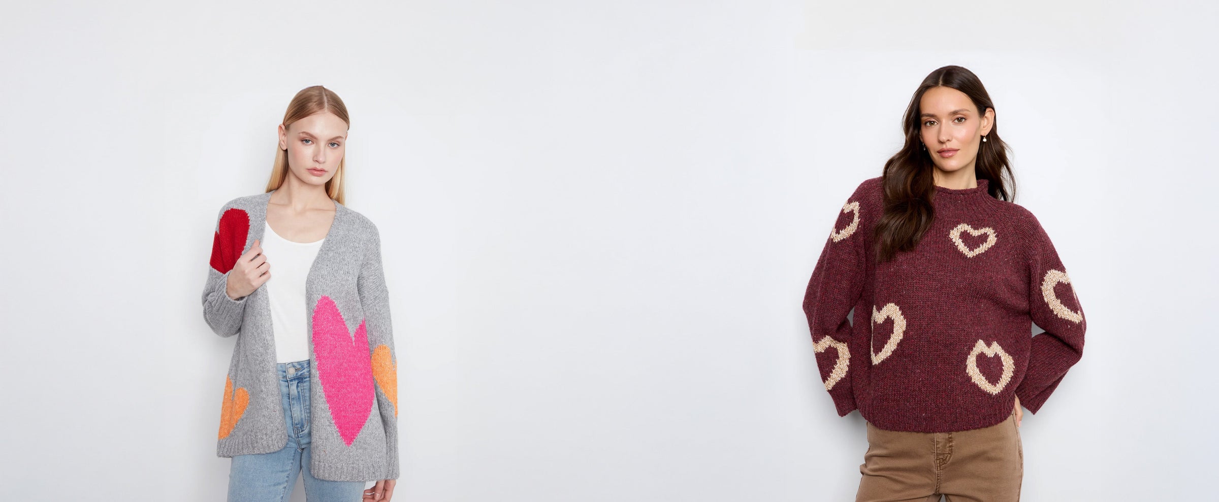 Two models standing against a plain white studio background, each wearing a knit sweater/cardigan with heart motifs. The model on the left wears a gray cardigan with pink and orange heart shapes over a white top and blue jeans, holding a red item.